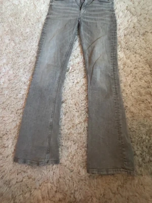 Ljusgrå bootcut jeans - Säljer ett par ljusgrå jeans med bootcut passform och klassisk femficksdesign. Jeansen har en rak midja och är tillverkade i denim, vilket ger en avslappnad och trendig look. Perfekta för dig som gillar en retrostil med utsvängda ben.