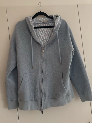 Ljusgrå hoodie med mönstrad huva - Säljer en ljusgrå hoodie med dragkedja och fickor framtill. Huvan har ett snyggt vitt och grått mönster på insidan samt dragsnören. Tillverkad i mjukt sweatshirtmaterial, perfekt för en avslappnad stil.