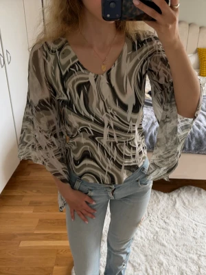 Mönstrad mesh topp med vida ärmar - Säljer en snygg mesh topp med abstrakt mönster i beige, svart och grå toner. Toppen har vida, genomskinliga långärmar och en tight passform som framhäver figuren. Perfekt för dig som vill sticka ut med en unik look.
