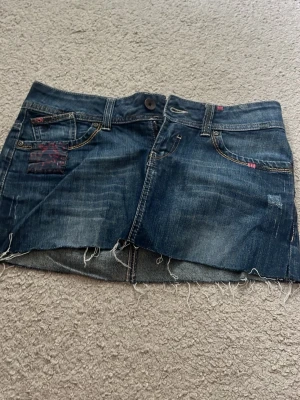 Mörkblå jeanskjol Espirit - Säljer en mörkblå jeanskjol i kort modell med rå, fransig kant nertill. Kjolen har klassiska fem fickor, knapp och dragkedja framtill samt slitna detaljer för en cool look. Perfekt till sneakers eller boots för en avslappnad stil.