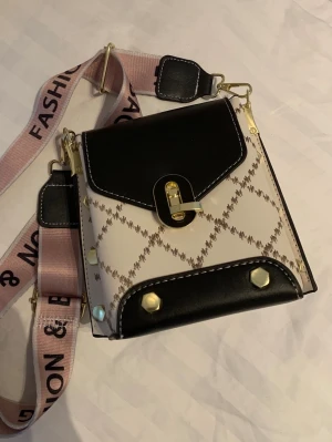 Beige och svart axelväska med mönster - Säljer en trendig axelväska i beige med brunt rutmönster och svarta detaljer. Väskan har guldfärgade metalldetaljer, ett lock med spänne och en bred rosa axelrem med text. 