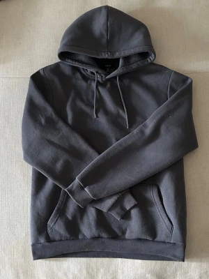 Mörkgrå hoodie från SMOG - En stilren mörkgrå hoodie från SMOG med klassisk huva och dragsnören. Hoodien har en känguruficka framtill och ribbade muddar vid ärmslut och nederkant. Perfekt för en avslappnad look och enkel att matcha med jeans eller joggers. Är ganska tjock och väldigt skön och komfortabel. 
