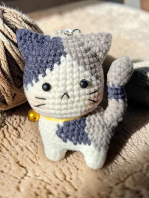 Virkad sur katt-nyckelring (handgjord)  - Handvirkad nyckelring i form av en katt med grå, mörk blå och vita partier. Katten har en liten gul bjällra runt halsen. Dockan virkade med omsorg och noggrann het. Nu är den redo att få en ny ägare.                          Storlek (ungefärlig)                    Huvudomkrets : 17.5 cm                            Höjd : 9 cm 