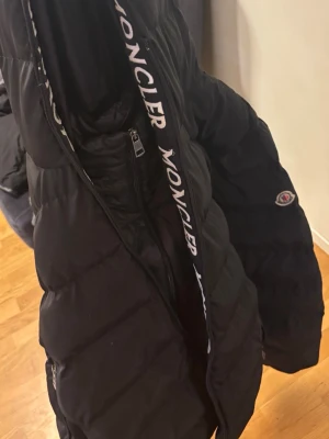 Moncler jacka  - Snygg svart dunjacka från Moncler med logotyp på ärmen och tydlig Moncler-text längs insidan av huvan. Jackan har dragkedja framtill och två fickor med dragkedja. Perfekt för kalla vinterdagar och riktigt trendig stil.