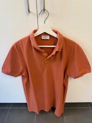 Pikétröja Rose & Born, stl L/52 - Snygg orange pikétröja från Rose & Born med klassisk krage och korta ärmar. Tillverkad i mjuk bomull som känns skön mot huden. Perfekt till jeans eller shorts för en avslappnad look.