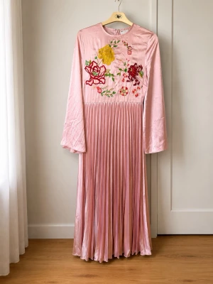 Lång rosa klänning med broderi - Elegant långklänning i ljusrosa satin med plisserad kjol och långa ärmar. Framtill finns vackra broderade blommor i rött, gult och grönt. Klänningen har rund halsringning och stängs med dragkedja i ryggen.