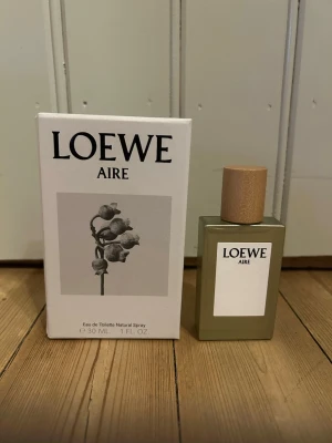 Loewe Aire Eau de Toilette 30 ml - Loewe Aire Eau de Toilette i 30 ml flaska. Parfymen kommer i en stilren, rektangulär glasflaska med träfärgat lock och vit etikett. Förpackningen är vit med minimalistisk design och en svartvit blomillustration. Perfekt för dig som gillar exklusiva dofter. Endast sprutat ett sprut så i princip helt ny!