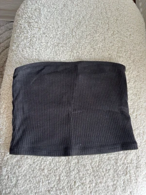 Svart ribbad tubtopp - Säljer en svart ribbad tubtopp i stretchigt material. Toppen är axelbandslös och har en enkel, stilren design som passar perfekt till många olika outfits. Perfekt för dig som gillar minimalistisk stil.