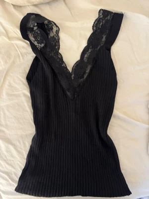 Svart ribbad linnetopp med spets - Snygg svart ribbad linnetopp med djup v-ringning och bred spetskant framtill. Toppen är ärmlös och har en figurnära passform som framhäver siluetten. Perfekt att styla med jeans eller kjol för en trendig look.