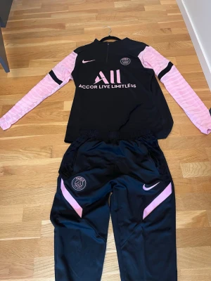 PSG träningsset svart/rosa Nike - Säljer ett PSG träningsset från Nike i svart med rosa detaljer. Tröjan har långa rosa ärmar, halv dragkedja och PSG-logga på bröstet. Byxorna är svarta med rosa detaljer och PSG-märke. Perfekt för fotbollsträning eller chill.