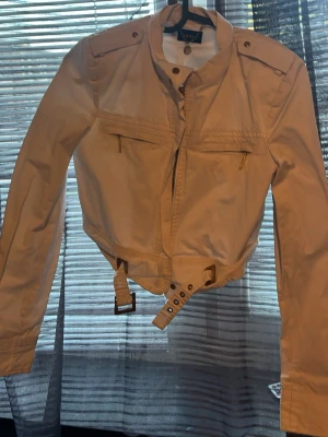 Beige bomberjacka med bälte - Snygg beige bomberjacka med bälte i midjan och dragkedja framtill. Jackan har långa ärmar, axelvaddar och två bröstfickor med dragkedja. Perfekt för dig som vill ha en stilren och trendig look.