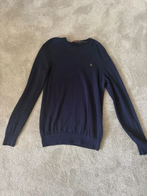 Mörkblå stickad tröja - Mörkblå stickad tröja med rund halsringning och långa ärmar. Tröjan har ribbade muddar vid ärmslut och nederkant samt en liten broderad detalj på bröstet. Perfekt för lager på lager och enkel att matcha med olika outfits.