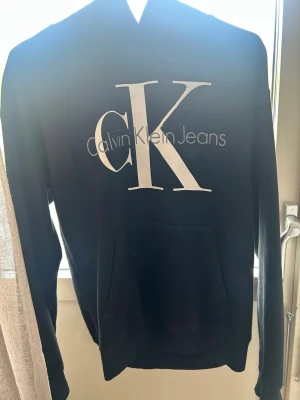 Svart hoodie från Calvin Klein Jeans - Snygg svart hoodie från Calvin Klein Jeans med stor vit CK-logga och text på bröstet. Klassisk huva baktill😍