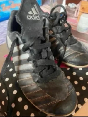 Svarta Adidas fotbollsskor med vita ränder - Säljer ett par klassiska svarta Adidas fotbollsskor med vita ränder på sidorna och snörning framtill. Skorna har en låg profil och slitstark gummisula med dobbar för bra grepp på planen. Perfekta för dig som vill ha stilrena och funktionella skor till match eller träning.Behöver tvättas 