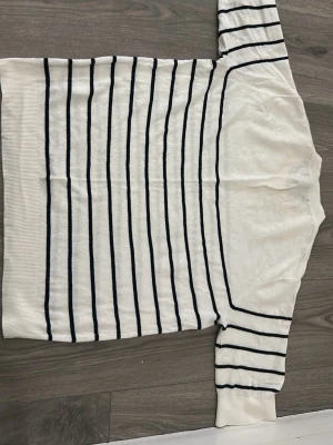 Vit stickad tröja med svarta ränder - Säljer en vit stickad tröja med tunna svart  ränder. Tröjan har rund halsringning, medium lång armar och ribbade muddar vid ärmslut och nederkant. Perfekt för en klassisk och stilren look.