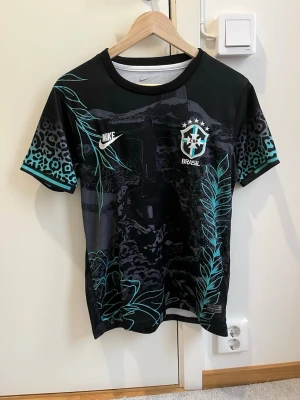 Nike Brasil svart matchtröja - Snygg svart matchtröja från Nike med Brasil-logga på bröstet. Tröjan har ett unikt mönster med turkosa blad och leoparddetaljer, samt vita loggor. Perfekt för dig som gillar fotboll och vill sticka ut på planen.