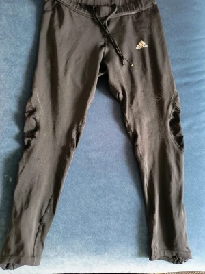 Svarta leggings från Adidas - Snygga svarta leggings från Adidas med klassisk logga på låret. De har en åtsittande passform, resår i midjan och dragsko för extra komfort. Tillverkade i mjukt och stretchigt material som passar perfekt till träning eller chill. Midjan 33cm och benen 75cm.
