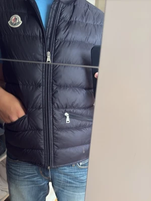 Svart dunväst från Moncler - Snygg svart dunväst från Moncler med klassisk quiltad design och dragkedja framtill. Västen har två fickor med dragkedja och Moncler-logga på bröstet. Perfekt att slänga över en hoodie eller tröja för extra värme.