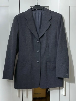 Mörkgrå klassisk kavaj - Stilren mörkgrå kavaj. Klassisk krage och helfodrad insida ger en snygg och tidlös look. Perfekt till både skjorta och t-shirt. Materialet ser ut att vara en blandning av polyester och viskos för en mjuk känsla.   Mått: längden är ca 78cm, axelbredden är ca 46cm och ärmlängden är ca 61cm
