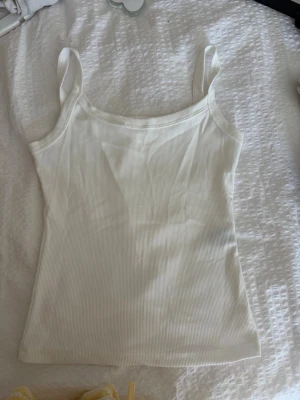 Vit ribbad linnetopp - Säljer en enkel och fräsch vit ribbad linnetopp med breda axelband. Toppen är ärmlös och har en tight passform som sitter snyggt på kroppen. Perfekt till sommaren och lätt att matcha med olika outfits.