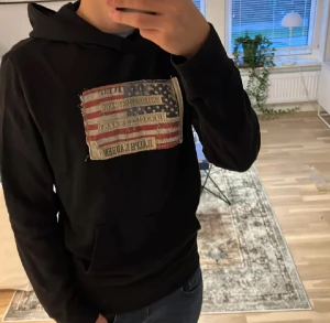 Svart hoodie från Ralph Lauren - Svart hoodie från Ralph Lauren med stor patch framtill i amerikansk flaggdesign och broderad logga. Tröjan har huva, känguruficka och långa ärmar. Perfekt för dig som gillar streetwear och vill ha något som sticker ut.väldigt efterfrågad.