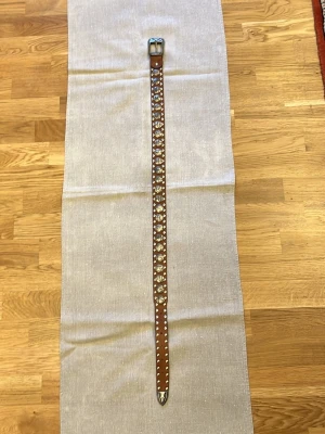 Brunt skärp med silverdetaljer 98cm - Snyggt brunt skärp i skinn med coola silverfärgade nitar och detaljer längs hela bandet. Spännet och spetsen har också silvertonade inslag som ger ett unikt uttryck. Perfekt accessoar för att lyfta din outfit.