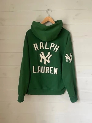 Grön Yankees hoodie Ralph Lauren - Grön hoodie med stor luva och dragsko från Ralph Lauren. Broderad text 'Yankees' framtill och 'Ralph Lauren' samt Yankees-logga på ryggen och ärmen. Klassisk känguruficka och ribbade muddar. Perfekt för dig som gillar sportig streetstyle.