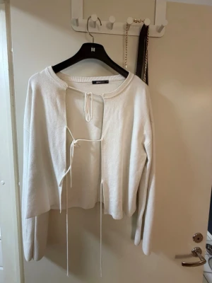 Vit stickad cardigan med knytband - Trendig vit stickad cardigan med öppen front och två knytband framtill. Långärmad modell med avslappnad passform och rund halsringning. Perfekt att styla över ett linne eller topp för en cool lager-på-lager-look.