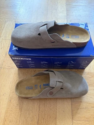 Birkenstock Boston tofflor i taupe - Säljer ett par klassiska Birkenstock Boston tofflor i taupe mocka. De har en justerbar rem med metallspänne och en bekväm korksula. Perfekta för dig som vill ha både stil och komfort hemma eller ute. Tidlös design som passar till det mesta.