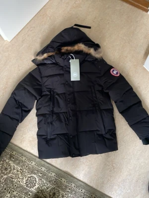 Svart dunjacka från Canada Goose - Säljer en svart dunjacka från Canada Goose med luva och avtagbar fuskpäls. Jackan har en klassisk patch på ärmen, dragkedja och knappar framtill samt flera fickor. Perfekt för kalla vinterdagar och riktigt varm.