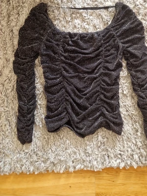 Svart glittrig långärmad topp - Säljer en svart glittrig topp med långa ärmar och rynkade detaljer framtill och på ärmarna. Toppen har en fyrkantig ringning och är figurnära i modellen. Perfekt för dig som vill sticka ut lite extra.