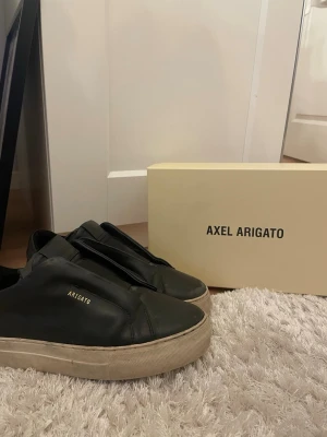 Svarta sneakers från Axel Arigato - Säljer ett par svarta sneakers från Axel Arigato i skinn med minimalistisk design och guldfärgad logga på sidan. Skorna har vit platt sula och rund tå. Medföljer originalkartong och dustbag.