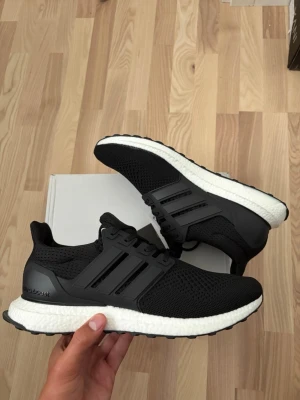 Svarta Adidas Ultraboost sneakers - Säljer ett par svarta Adidas Ultraboost sneakers med vit sula och klassiska tre ränder på sidan. Ovandelen är i ventilerande mesh och sulan är i mjukt, stötdämpande material för extra komfort. Perfekta för dig som vill ha både stil och funktion.