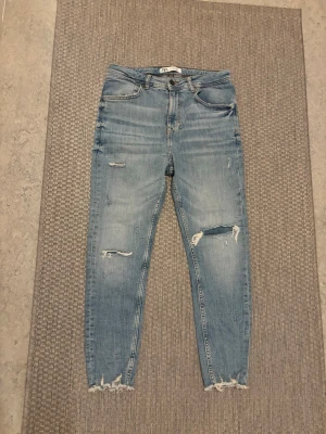 Ljusa slitna jeans från ZARA - Säljer ett par ljusblå jeans från ZARA med trendiga slitningar på benen och fransiga benslut. Jeansen har klassisk femficksmodell och normal midja. Perfekta för en avslappnad och cool stil.