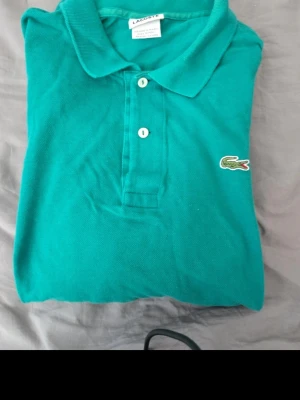 Grön pikétröja från Lacoste - Snygg grön pikétröja från Lacoste med klassisk krage och två knappar framtill. Tröjan har det ikoniska krokodilmärket broderat på bröstet och är tillverkad i bomull. Perfekt för en stilren och sportig look.
