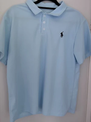 Ljusblå pikétröja från Polo Ralph Lauren - Klassisk ljusblå pikétröja från Polo Ralph Lauren med broderad logga på bröstet. Tröjan har korta ärmar, krage och knappar framtill. Perfekt för en clean och stilren look.