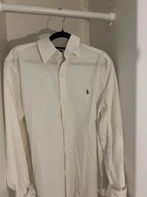 Vit skjorta med broderad logga - Klassisk vit skjorta i bomull med lång ärm och knappar framtill. Skjortan har en broderad logga på bröstet och en stilren krage. Perfekt till många olika looks och enkel att matcha.