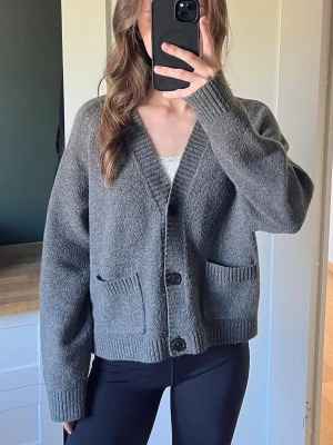 Grå oversized kofta med fickor - Mysig grå kofta i oversized modell med två stora fickor framtill och knäppning med svarta knappar. Koftan har ribbade muddar vid ärmslut och nederkant, perfekt för lager-på-lager. Passar dig som gillar en avslappnad och trendig stil.