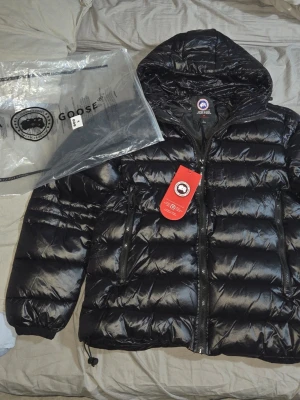 Svart dunjacka från Canada Goose - Snygg svart dunjacka från Canada Goose med glansig finish och huva. Jackan har två dragkedjeförsedda fickor framtill och justerbar nederkant. Perfekt för kalla vinterdagar och riktigt varm tack vare dunfyllningen.