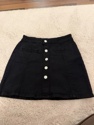 Svart kortkjol med knappar framtill - Snygg svart kortkjol i denim med fem silverfärgade knappar framtill. Kjolen har två dekorativa fickor och en lätt A-linjeformad siluett. Perfekt att matcha med en enkel topp eller stickad tröja.