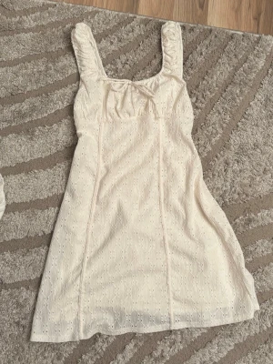 Vit kort klänning med broderade detaljer - Söt vit kort klänning med breda axelband och broderade hål för en romantisk look. Klänningen har en liten rosett framtill och är tillverkad i ett lätt bomullstyg, perfekt för varma dagar.