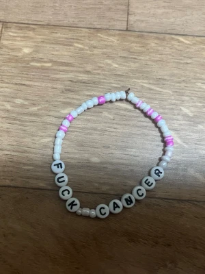 Pärlarmband med text Fuck Cancer - Säljer ett handgjort armband med vita och rosa pärlor samt bokstavspärlor som stavar 'FUCK CANCER'. Armbandet är elastiskt och har en ungdomlig, personlig stil. Perfekt för dig som vill visa stöd och samtidigt bära något unikt. Bokstaven N är lite förstörd men ändå användbar (se bild) 