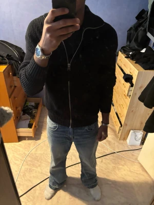 Svart stickad ziptröja - Säljer en svart stickad ziptröja med hög krage och grov ribbstickning. Tröjan har dragkedja framtill och långa ärmar, perfekt att slänga på över en t-shirt för en clean look.