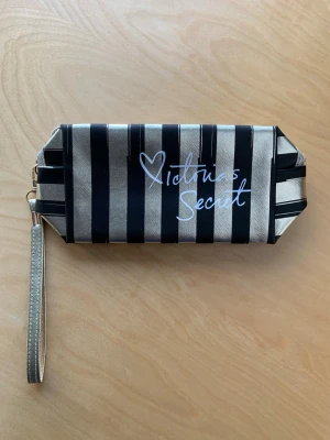 Randig plånbok Victoria's Secret - Snygg plånbok från Victoria's Secret med svarta och guldiga ränder. Plånboken har ett handledsband i matchande färg och loggan i vitt framtill. Tillverkad i ett glansigt tygmaterial och har en modern, rektangulär form.