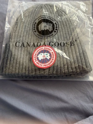 Grå ribbstickad mössa Canada Goose - Säljer en grå ribbstickad mössa från Canada Goose med klassisk uppvikt kant och det ikoniska runda märket framtill. Mössan är tillverkad i mjukt material och har en stilren look som passar perfekt till kalla dagar.