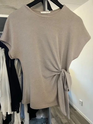 Beige topp med knyt i sidan - Säljer en beige topp med korta ärmar och snygg knytning i sidan. Toppen har en rund halsringning och är tillverkad i ett mjukt, stickat material. Perfekt för dig som gillar stilrena plagg med en twist.