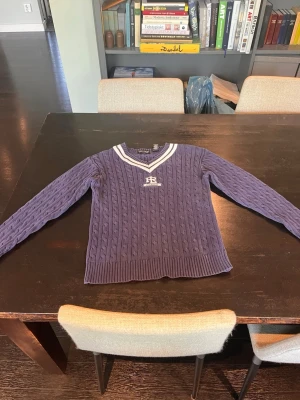 Blå kabelstickad tröja Polo Ralph Lauren - Kabelstickad blå tröja från Polo Ralph Lauren i 100% bomull. V-ringad med vita ränder runt halsen och broderad logga på bröstet. Långa ärmar och ribbstickade muddar. Perfekt för en preppy look.