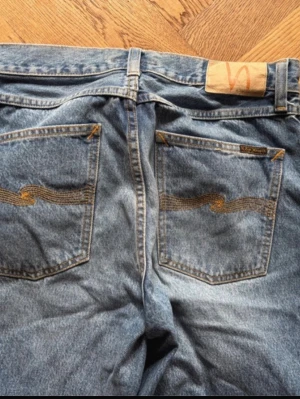 Blå raka jeans från Nudie Jeans - Säljer ett par klassiska blå jeans från Nudie Jeans med raka ben och hög midja. Jeansen har tydliga kontrastsömmar och ikoniska broderade bakfickor. Tillverkade i slitstark denim med vintagekänsla och femficksmodell. Perfekta för en avslappnad och trendig look. 30/34