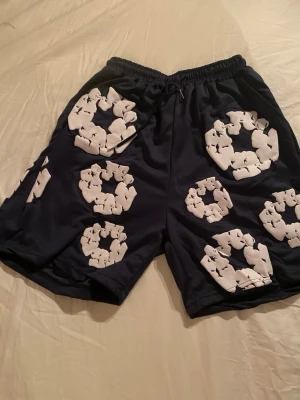 Denim Tears shorts - Säljer ett par marinblå shorts med elastisk midja och snörning. Shortsen har ett unikt vitt grafiskt mönster över hela plagget och är tillverkade i ett mjukt syntetmaterial som passar perfekt till sommaren.