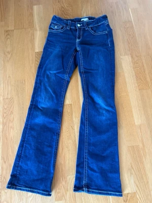 Mörkblå bootcut jeans med kontrastsömmar - Snygga mörkblå bootcut jeans med vita kontrastsömmar och klassiska fem fickor. Jeansen har utsvängda ben och detaljerade bakfickor med knapp. Inte använda många gånger på grund av att jag beställt fel storlek och inte använda på ett tag därför skrynkliga.